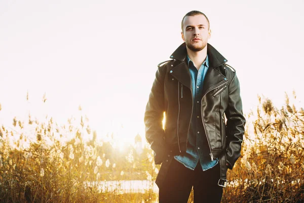 Handsome young man in leather jacket in the autumn park on a sunset. Seasonal fashion.