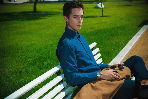 Handsome imposing young man is sitting on a bench in a park. Fashion shot. Business style. 
