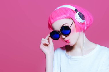 Trendy kız pembe saçlı güneş gözlüğü takıyor kulaklıklar müzik hoşlanır. Pembe bir arka plan. Gençlik stil, eğlence.