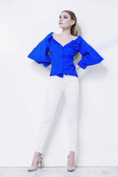 Attractive young woman in a blue blouse and white pants stands by a white wall. Beauty, fashion concept. Full length portrait.