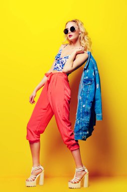 Çekici genç kadın sarı arka plan üzerinde parlak giysiler ve güneş gözlüğü takıyor. Parlak stil, moda. Optik tarzı. Tam uzunlukta portre.