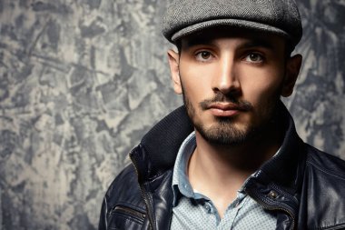 Grunge arka plan üzerinde bir kap ve deri ceketli yakışıklı bir adam portresi. Stüdyo vurdu. Erkek güzelliği, moda. Erkek berber.