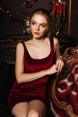Kadife kırmızı elbiseli güzel genç kadın bir çift koltukta oturuyor. Klasik vintage iç. Güzellik, moda.