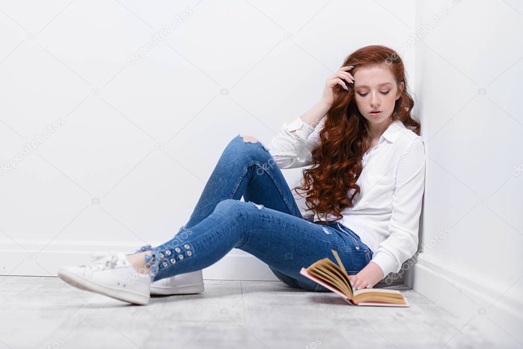 Beautiful red-haired teen girl wearing in jeans and blouse reading a book. Beauty, fashion.