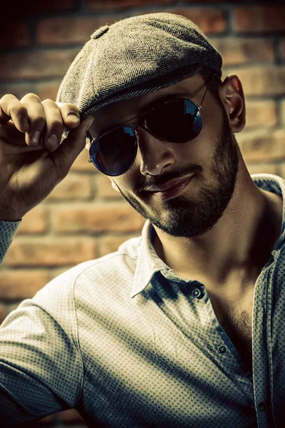 Portrait of a handsome man in a cap and sunglasses over brick wall background. Men's beauty, fashion. Men's barbershop.
