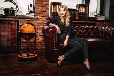 Çekici genç kadın deri kanepe tuğla duvarlı bir odada poz. Loft tarzı iç. Güzellik, moda. Akşam makyaj ve saç.