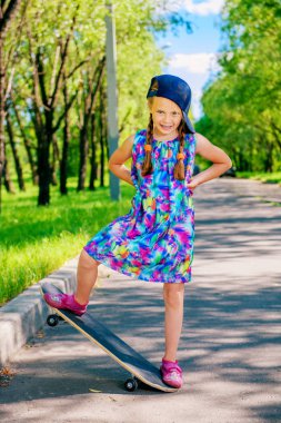 Cheerful little girl riding on a skateboard in the park. Outdoor activity. Summer holidays. clipart