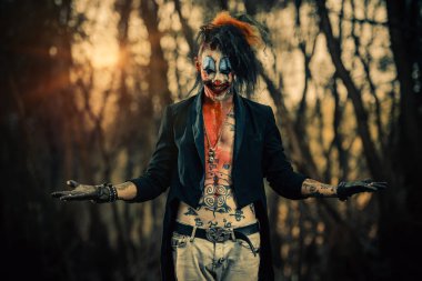 Crazy evil clown man is standing in the forest. Halloween. Horror, thriller movie. clipart