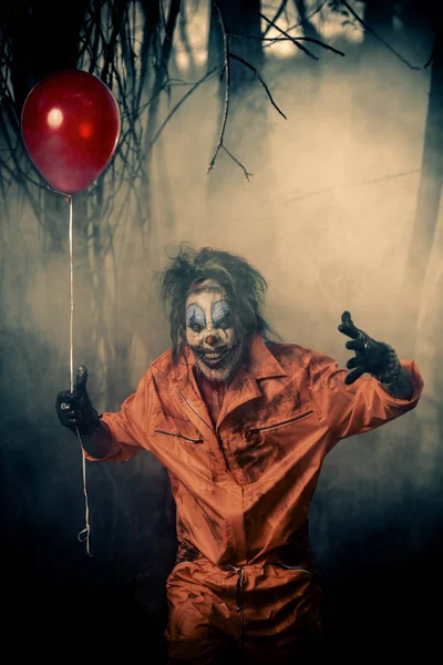 Scary man clown stained in blood in a night forest with a balloon. Male zombie clown. Halloween. Horror.