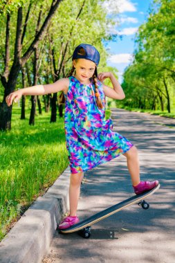 Cheerful little girl riding on a skateboard in the park. Outdoor activity. Summer holidays. clipart