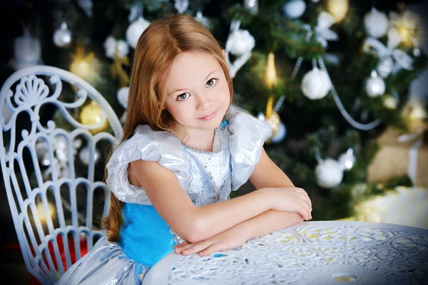 Cute six-year-old girl sits at a table near the Christmas tree. Classic luxurious interior decorated for Christmas. Merry Christmas and Happy New Year.