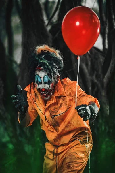 Scary man clown stained in blood in a night forest with a balloon. Male zombie clown. Halloween. Horror.