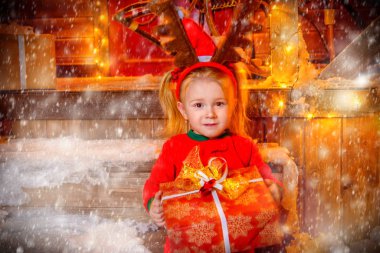 Oldukça üç yıl yaşlı kız Noel için bir geyik dekore evinin yakınında oturuyor ve bir hediye kutusu tutarak baştan boynuzlarıyla uygun. Mutlu Noeller ve mutlu yeni yıl.