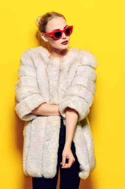 Çok güzel sarışın kadın lüks kürk ceket ve pin-up güneş gözlüğü içinde poz. Sarı arka plan. Moda, güzellik. Stüdyo vurdu.