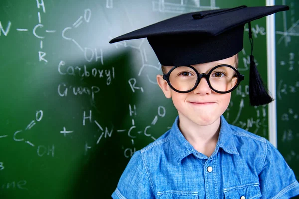 A portrait of a young boy in glasses posing over the blackboard at school. Education, knowledge.