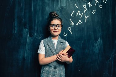 Bir kitap ile bir kara tahta üzerinde duran bir sevimli kız portresi. Okul, eğitim.