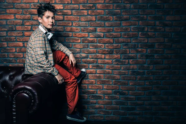 A full length portrait of a trendy boy wearing casual clothes sitting on the sofa in the interior. Fashion for male teenagers.