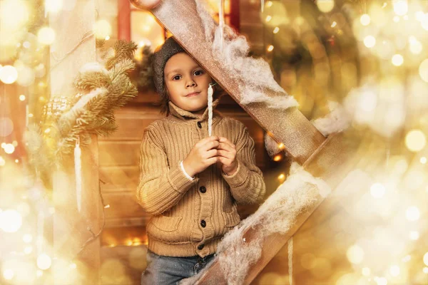 A little boy sitting on the porch of the house decorated for Christmas. Miracle time. Merry Christmas and Happy New Year.