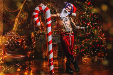 Bad Santa concept. Nude tattooed Santa with purple dreads holding a great Christmas Lollipop  and is demonstrating antisocial behavior, rudeness and aggression in luxury apartments. clipart