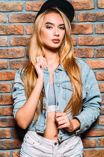 Uzun sarı saçlı kot giyen serin modern kız giysi ve beyzbol kap ayakta duvara tarafından. Hippi tarzı. Gençlik moda.