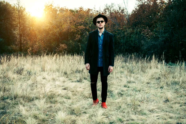 Sonbahar moda. Yakışıklı erkek model siyah ceket, şapka ve güneş gözlüğü açık poz. Sonbahar doğa.