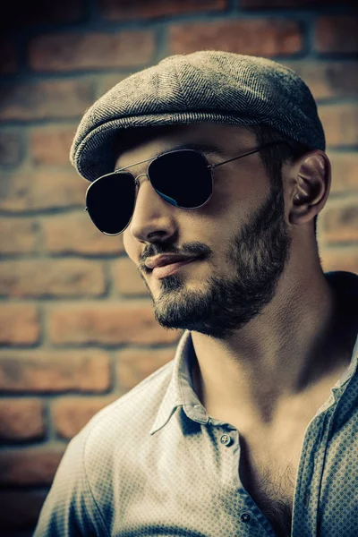 Portrait of a handsome man in a cap and sunglasses over brick wall background. Men\'s beauty, fashion. Men\'s barbershop.