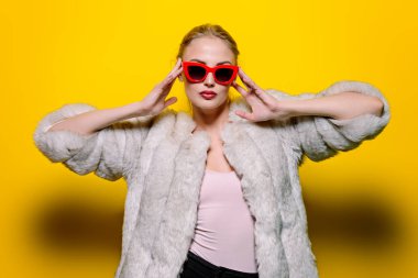 Çok güzel sarışın kadın lüks kürk ceket ve pin-up güneş gözlüğü içinde poz. Sarı arka plan. Moda, güzellik. Stüdyo vurdu.