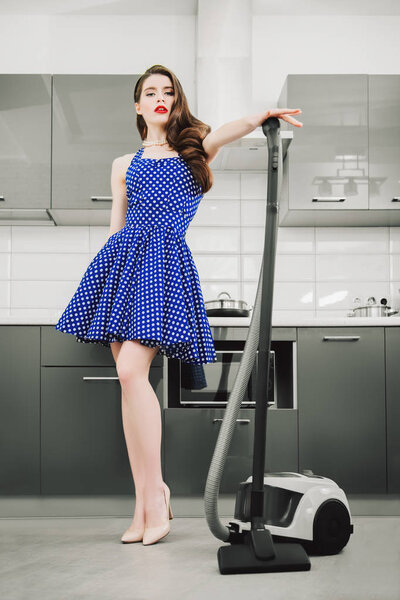 Beautiful young housewife wearing in a blue dress with white dots in the kitchen. Pin-up style. Fashion home shot. Full length portrait. 