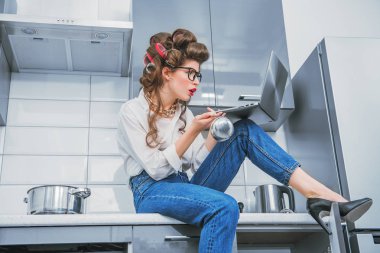 Güzel bir kız ile bir dizüstü bilgisayar ile saçında curlers günlük giysiler içinde poz portresi. Moda ev atış. Pin-up tarzı.