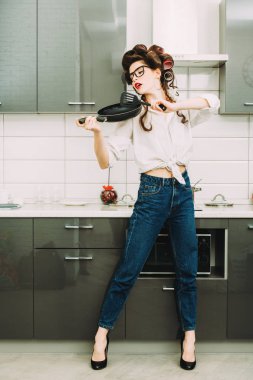 Günlük giysiler ve curlers çekici bir kız mutfakta bir tava ile ayarlamak anlamına gelir. Moda ev atış. Tam uzunlukta portre. 