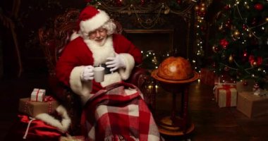 Noel Baba bir battaniye ile kaplı bir deri sandalyede oturuyor, Noel için dekore edilmiş bir odada bir fincan sıcak bir içecek içiyor. Noel ve Yeni Yıl zamanı.