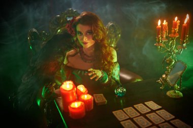 Güzel kızıl saçlı bir falcı siyah bir kuzgunla konuşuyor. Sihir ve büyü atmosferi, sihirli kristal küre ve tarot kartları..
