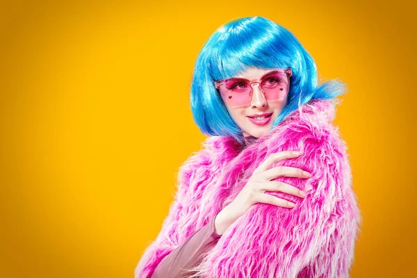 Portrait of an attractive party girl with bright pink makeup and blue wig wearing pink fur coat and sunglasses on a yellow background. Make-up and cosmetics, hairstyle. Fashion girl. Copy space.