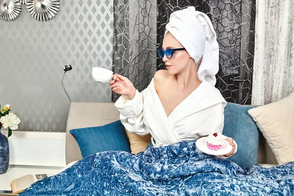 Glamorous young woman in elegant sunglasses, a white bathrobe and with a towel around her hair after a shower lies in her cozy bed, drinks coffee and eats a cake. Luxurious lifestyle.