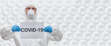 COVID-19 concept. A doctor in a protective suit holds a sign COVID-19 on a white background. Copy space. clipart