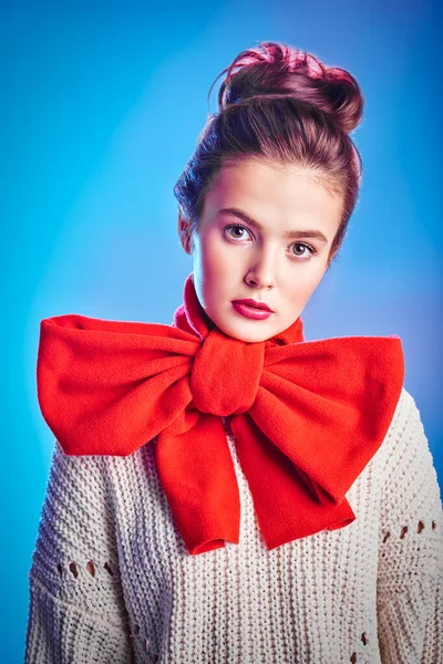 Portrait of an elegant beautiful girl in a white sweater and with a big red bow on her neck against a soft pink and blue light. Beauty, fashion concept. Winter style.