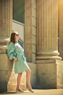 Şehir caddesinde duran zarif yaz elbiseli ve güneş gözlüklü zarif bir genç kadının tam boy portresi. Yaz modası.