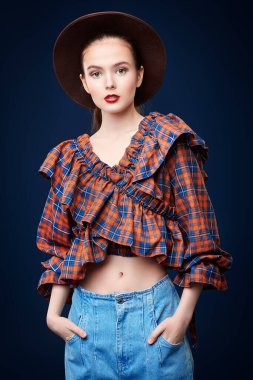 Moda fotoğrafı. Kot pantolon, kareli bluz ve kahverengi şapkayla poz veren güzel bir kız modeli portresi. Country, batı tarzı. Yeni yaz koleksiyonu.