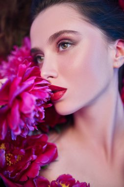 Güzellik ve çiçekler. Şakayık çiçeklerin arasında uzanan parlak makyajlı güzel esmer bir kadının yakın plan portresi. Makyaj malzemeleri, makyaj malzemeleri. Parfüm.