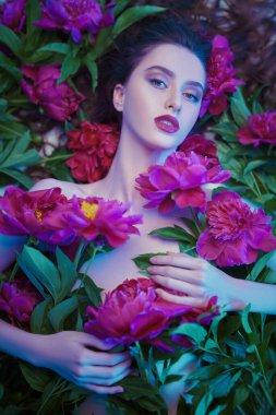 Güzellik ve çiçek konsepti. Şakayık çiçeklerin arasında uzanan parlak makyajlı güzel esmer bir kadının portresi. Makyaj malzemeleri, makyaj malzemeleri. Parfüm.