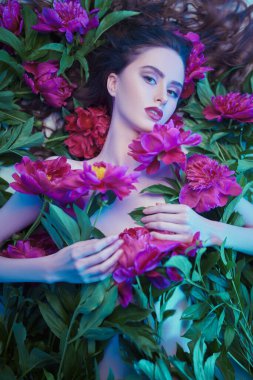 Güzellik ve çiçek konsepti. Şakayık çiçeklerin arasında uzanan parlak makyajlı güzel esmer bir kadının portresi. Makyaj malzemeleri, makyaj malzemeleri. Parfüm.