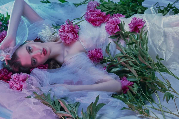 Şeffaf beyaz elbiseli çekici esmer kadın taze şakayık çiçeklerinin arasında yatıyor. Güzellik, moda portresi. Makyaj ve kozmetik.