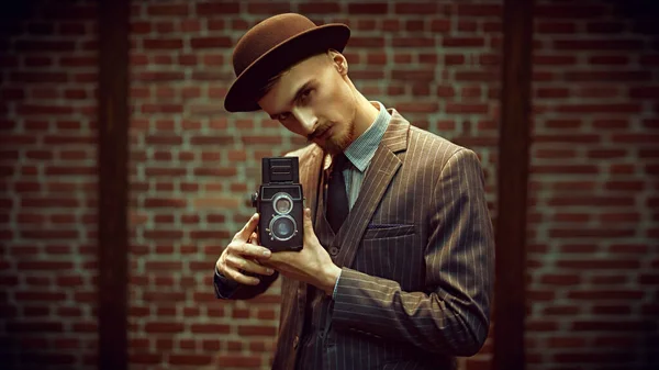 Retro concept. Handsome photographer man in an elegant suit is working with his old camera on a brick wall background.