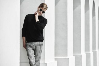 Moda fotoğrafı. Siyah kazaklı, ekose pantolonlu ve siyah gözlüklü yakışıklı genç adam eski kasabadaki beyaz sütunların yanında poz veriyor. Erkek stili, güzellik.