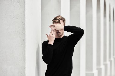 Sanat portresi. Siyah kazaklı yakışıklı genç adam eski kasabadaki beyaz sütunların yanında poz veriyor, yüzünü elleriyle örtüyor. Erkek stili, güzellik. Moda fotoğrafı.. 