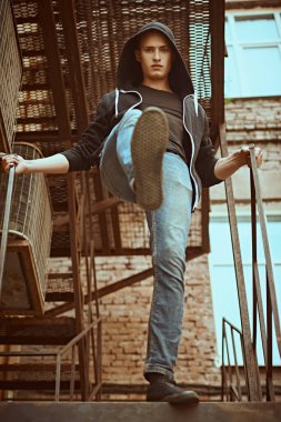 Saldırgan genç adam eski demir bir merdivende duruyor, kameraya bakıyor ve kameraya doğru bir adım atıyor. Kentsel grunge tarzı.