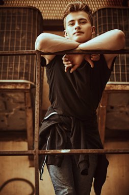 Sıradan kıyafetler içindeki yakışıklı genç adam metal endüstriyel bir merdivende duruyor ve hafifçe gülümsüyor. Şehirli grunge stili. Erkek modası.