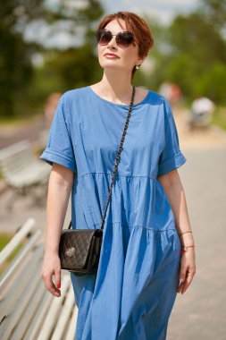 Zarif güneş gözlüklü ve uzun mavi elbiseli mutlu orta yaşlı kadın yaz parkında dinleniyor..