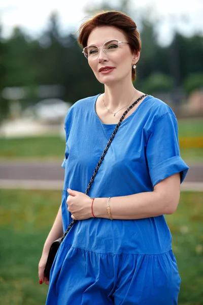 Happy middle-aged woman in elegant glasses and long blue dress having a rest in the summer park.