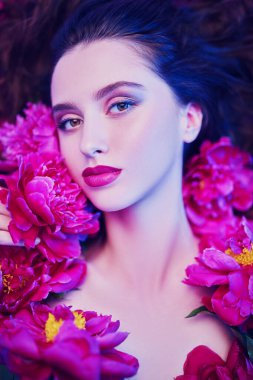 Güzellik ve çiçek konsepti. Şakayık çiçeklerin arasında uzanan parlak makyajlı güzel esmer bir kadının portresi. Makyaj malzemeleri, makyaj malzemeleri. Parfüm.